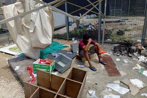 A man sits with his hand on his head, surrounding by trash and debris.