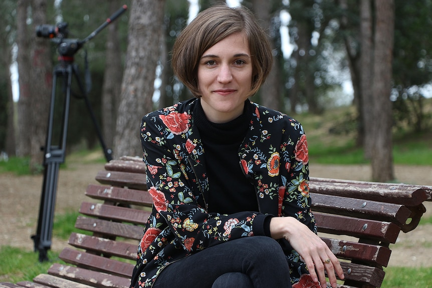 Colour mid-shot photo of director Carla Simon sitting cross-legged on a bench in front of tall trees and a camera on a tripod.