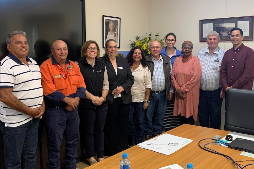 Indigenous elders and local government councillors stand around a table in a board room, smiling to the camera.