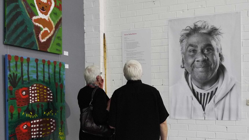 Two people standing in front of a black-and-white photograph of artist Trevor 'Turbo' Brown at an exhibition.