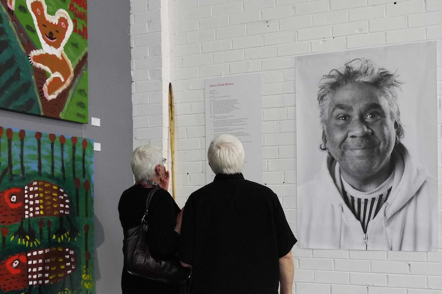 Two people standing in front of a black-and-white photograph of artist Trevor 'Turbo' Brown at an exhibition.