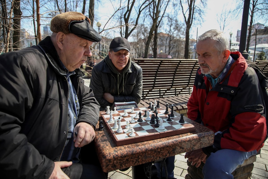 Three white men in winter clothes sit around a chess board in a park.