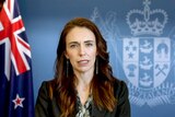 New Zealand PM Jacinda Ardern speaks in front of a New Zealand flag and coat of arms.