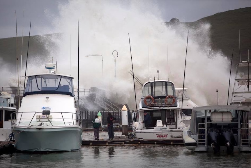 Waves crash over Coffs Harbour breakwall onto yachts moored in the marina.