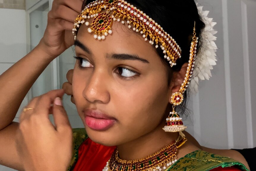 A young brown girl looks into the mirror as she adjusts intricately beaded jewellery around her face and ears. She is proud.