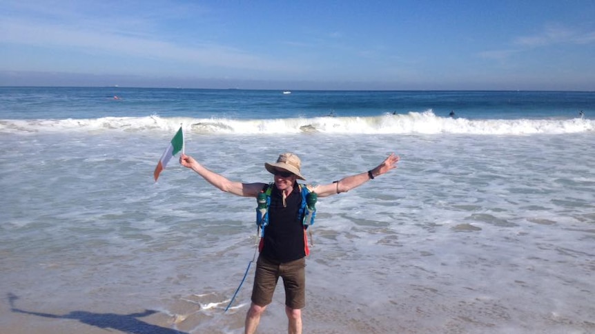 Tony Mangan stands on the beach in a backpack, holding an Irish flag.