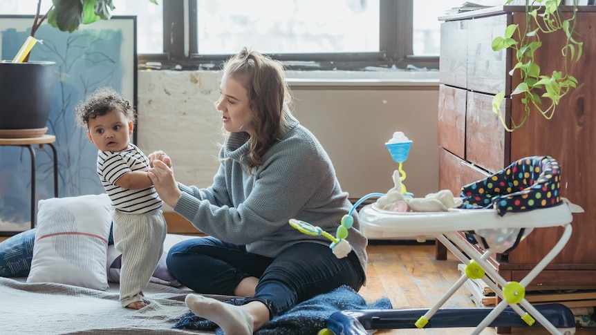 A woman sits on the floor of her home with her young toddler. 