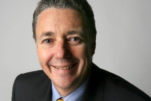 A headshot of Brett McCarthy wearing a suit and yellow tie.