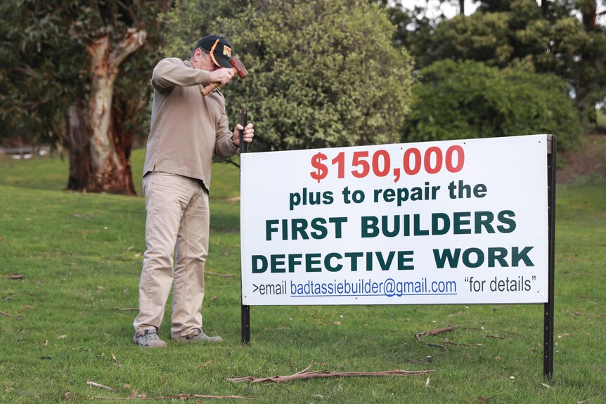 A man hammers in a sign saying, "$150,000 plus to repair the first builders defective work".