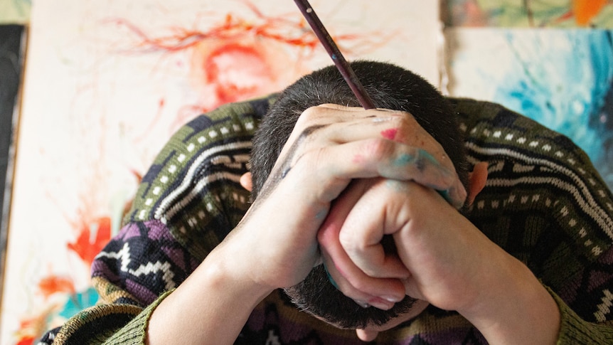 A white man holding a paintbrush slumps his head into his paint-splattered hands on a desk.