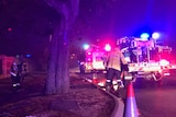 Two fire trucks with their lights on parked on the side of a road at night, with two firefighters running a hose.