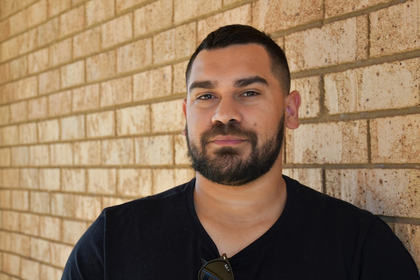 A bearded, dark-haired Aboriginal man wearing a dark T-shirt leans against a brick wall and smiles at the camera