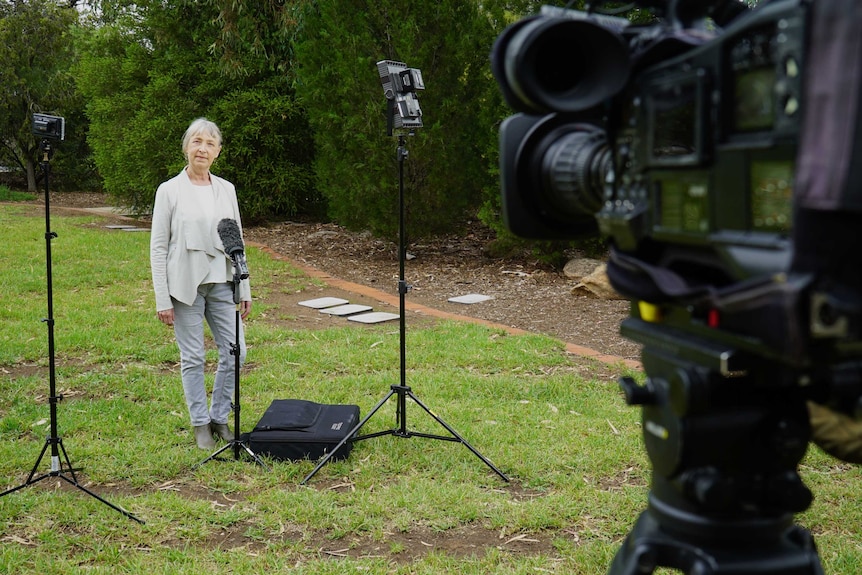 Woman standing in garden in front of mic on stand and lights on stands with camera a couple of metres away.