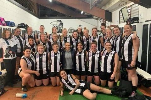 A group of women wearing black and white stripped AFL uniforms poses for a photo in the change rooms