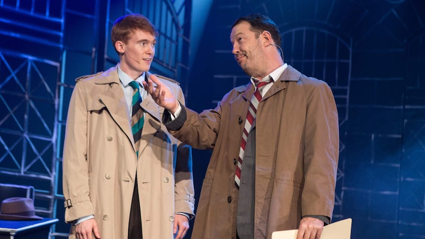 two men in 1940s detective costumes on a blue-lit stage, with wrought-iron-style scaffolding behind them.