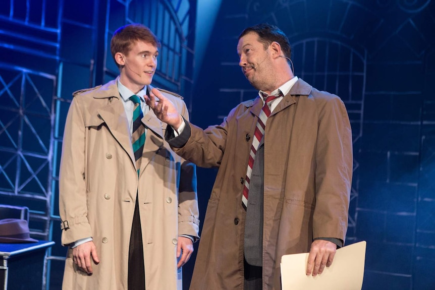two men in 1940s detective costumes on a blue-lit stage, with wrought-iron-style scaffolding behind them.