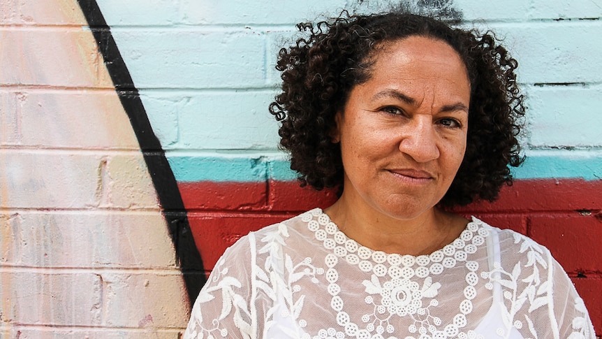 A woman with black curly hair and a white lace top stands in front of a wall