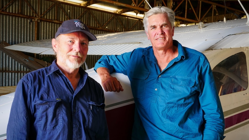 Two men stand in front of a light aircraft in a shed