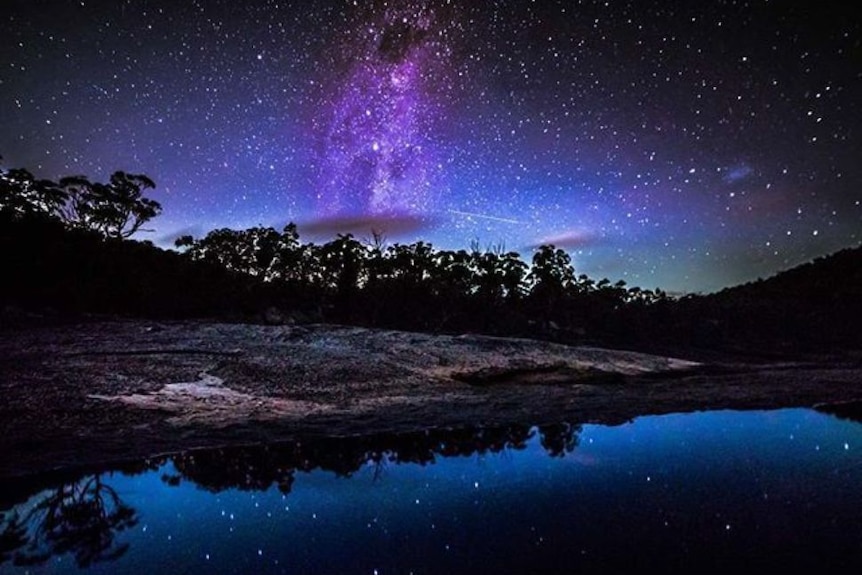 The Milky Way and stars  reflected in a lake, with trees on the horizon, at Girraween National Park in NSW.