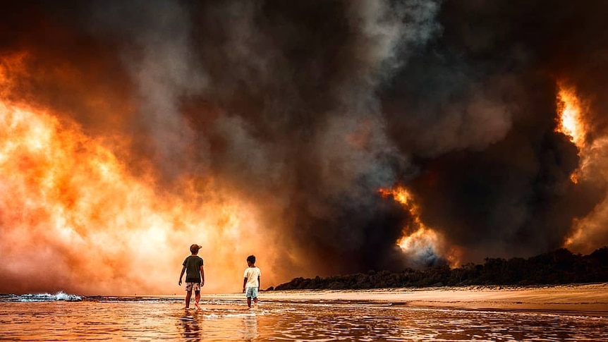 Two children stand on a beach. a cloud of red smoke obscures land from the background