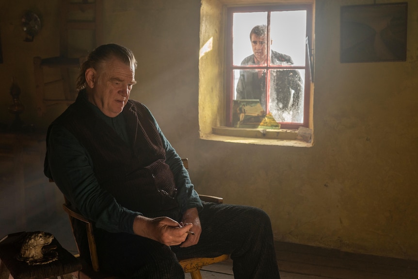 A man sits in a bleak-looking house smoking a cigarette as another, forlorn-looking man peers in at him through a window.