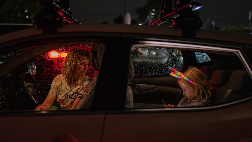 A woman sitting in the driver's seat of a car at night looks over her shoulder at a young girl in the back wearing bunny ears.