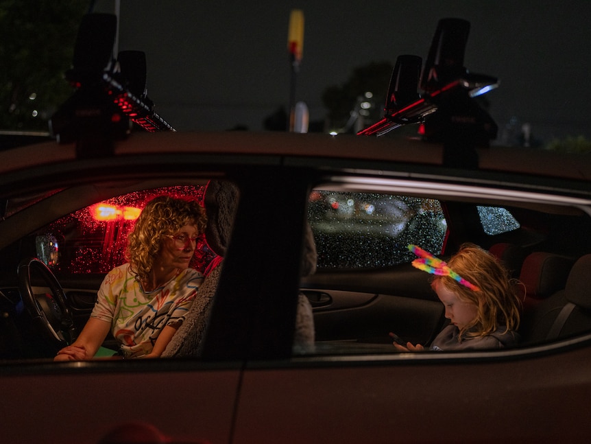 A woman sitting in the driver's seat of a car at night looks over her shoulder at a young girl in the back wearing bunny ears.