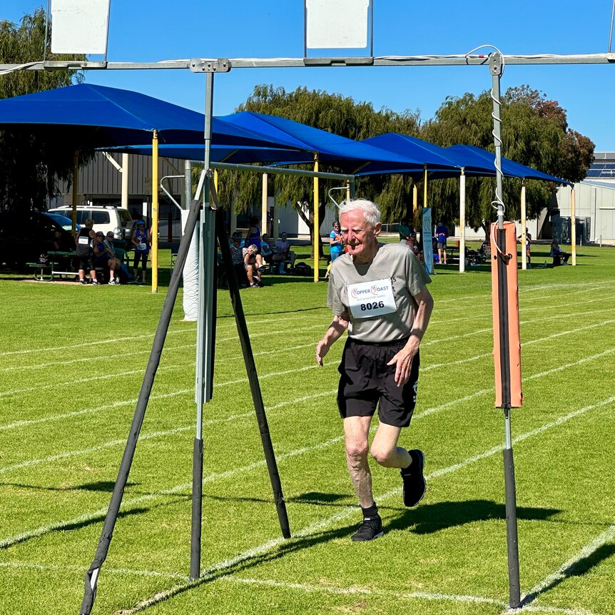 an elderly man in a polo top and shorts crosses the finishing line at a race on green grass