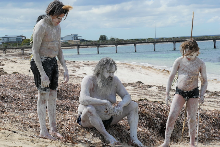 A man with long hair and two boys, covered in white ochre sit on a beach 