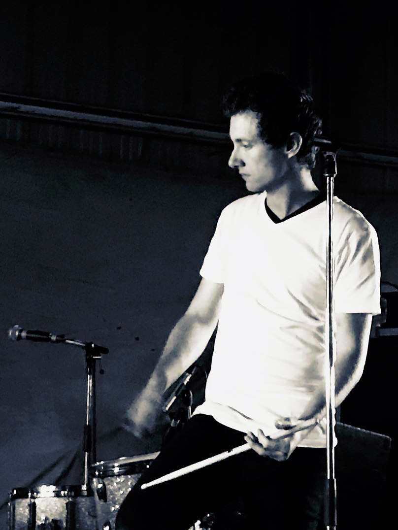 A young man with short brown hair and a white t-shirt plays drums near microphones.
