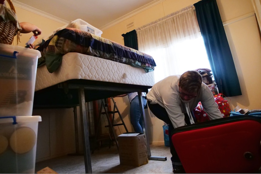 woman bends over to pick something off the ground. Large bed raised on a tall steel frame behind her