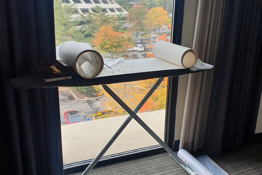 An ironing board with a rolled up piece of scroll with botanic pictures in front of a window.