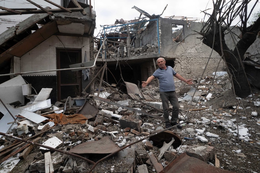 A man holds his hands out wide as he gestures while standing in the centre of a home destroyed during a shelling