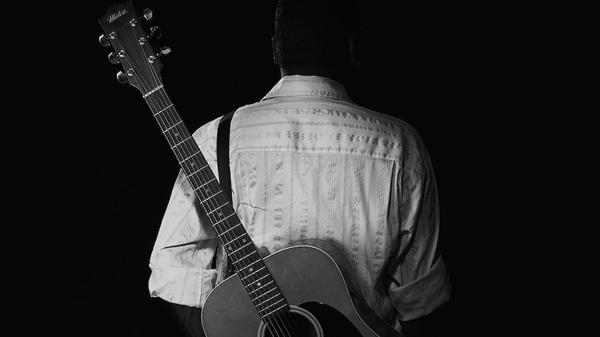 A photography of Gurrumul from behind, with a guitar slung diagonally across his back.