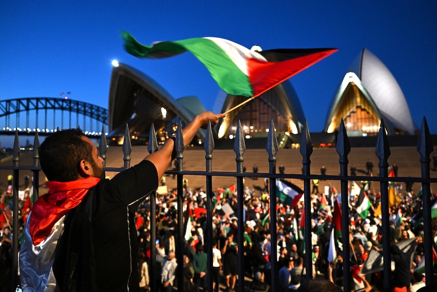 Man stands above Opera House forecourt on dusk waving Palestinian flag with sea of protestors below