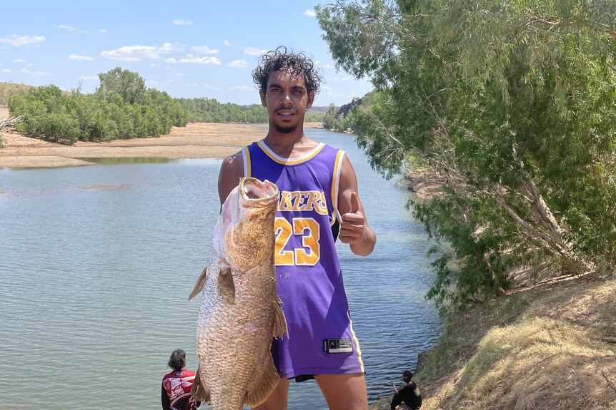 A young man standing on the bank of a river, holding a large fish with his thumb up.