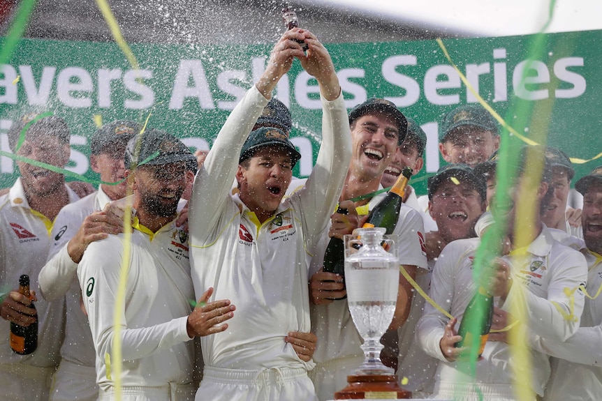 Australia captain Tim Paine lifts the Ashes Urn as his Australian teammates smile, laugh and spray champagne. Streamers fly.