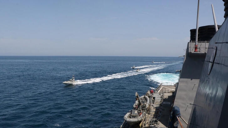 The edge of a US navy ship with Iranian navy vessel and a few crossing ships shown in the sea.