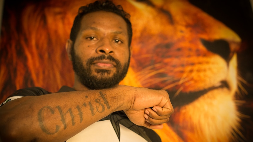 An Indigenous man stands in front of a large portrait of a lion, showing a tattoo on his arm that reads 'Christ'.