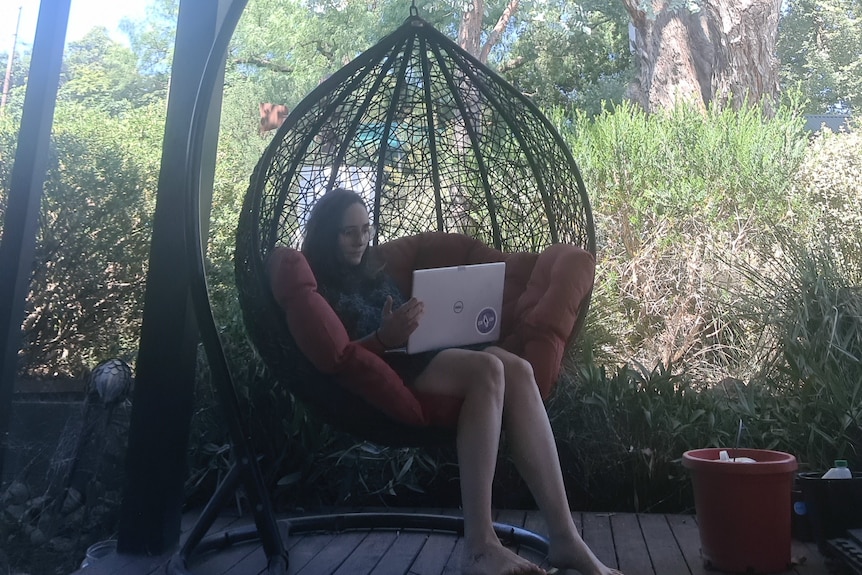 A young woman sits looking at a laptop in a hanging outside chair.