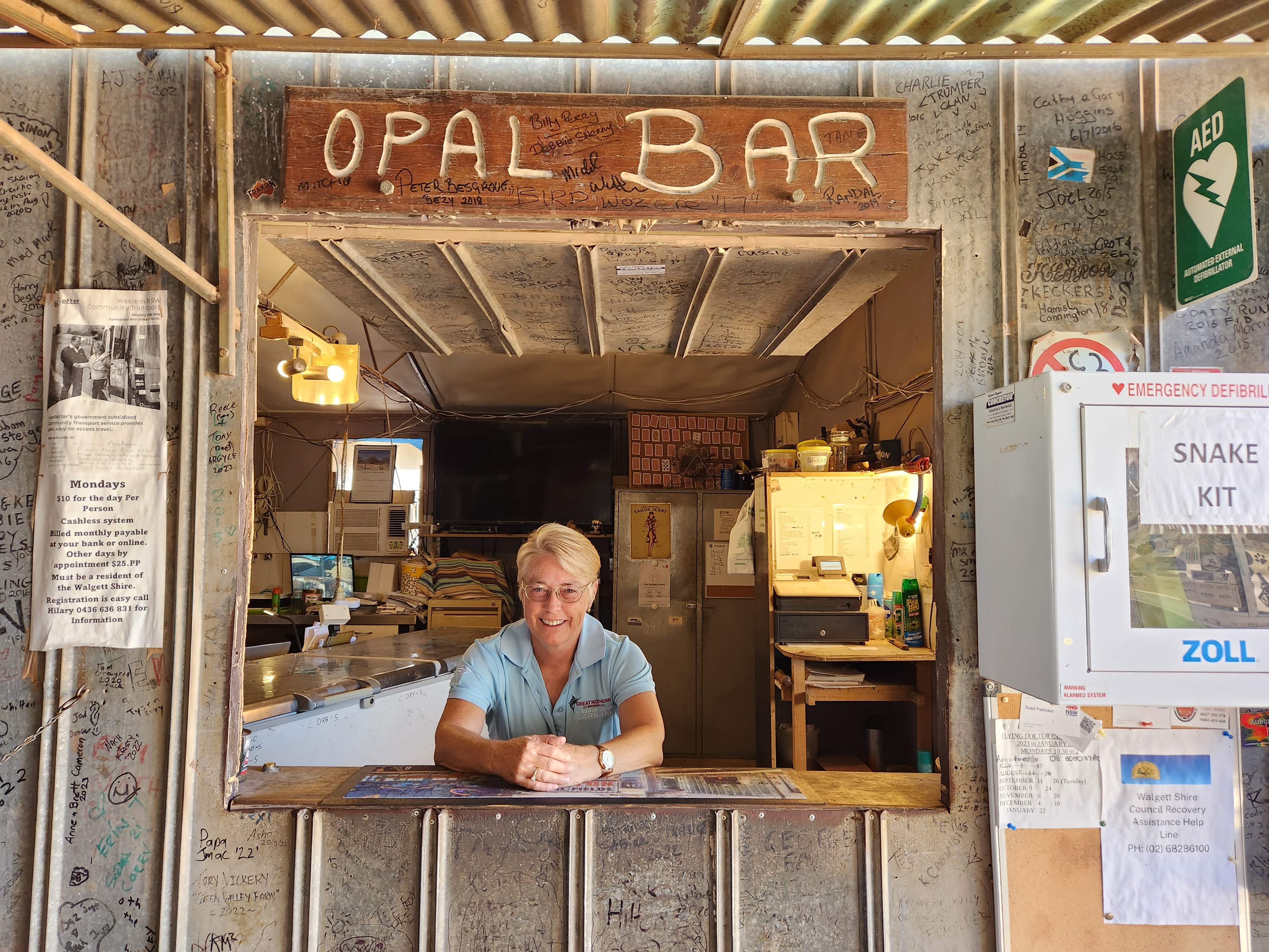A smiling, middle-aged woman leans on a bar beneath a sign that says "Opal Bar".