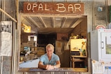 A smiling, middle-aged woman leans on a bar beneath a sign that says "Opal Bar".