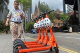 Brendon Donohue, who is blind and uses a cane, stands in front of line of e-scooters.