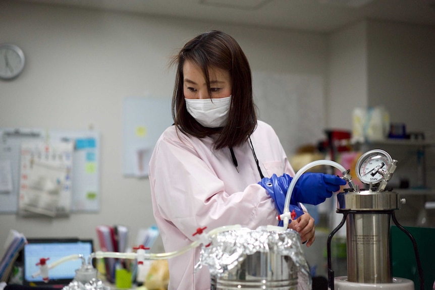 A Japanese woman in a face mask and gloves measuring radiation in a lab.