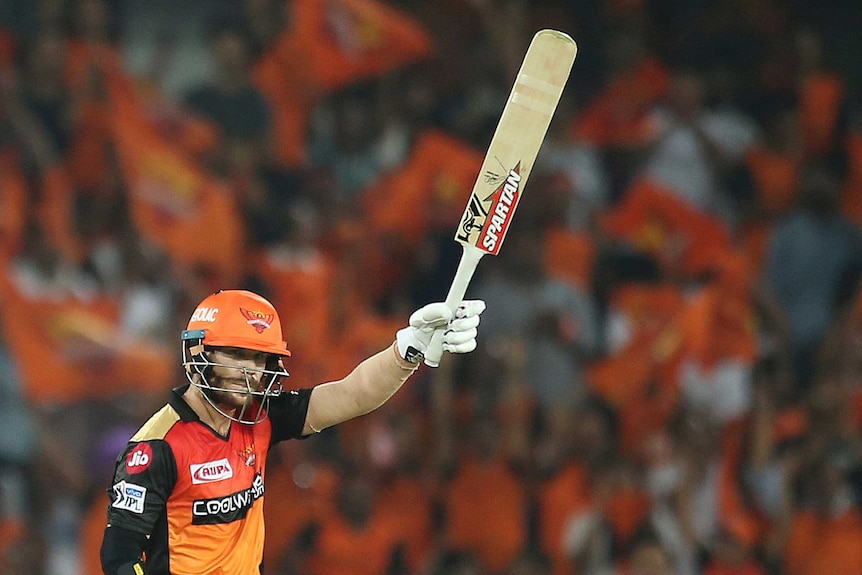 David Warner raises his bat above his head in an orange kit and orange helmet with orange flags being waved in the background