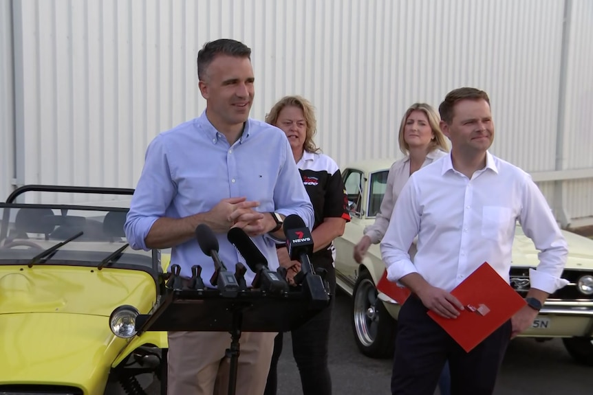 Two men in front and two women behind with cars