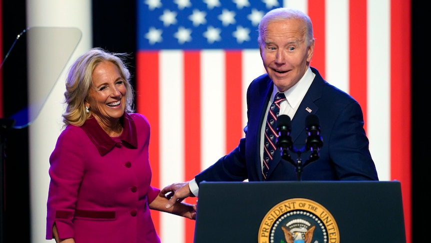 Jill Biden and Joen Biden laugh while standing at a podium in front of a large American flag