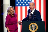Jill Biden and Joen Biden laugh while standing at a podium in front of a large American flag