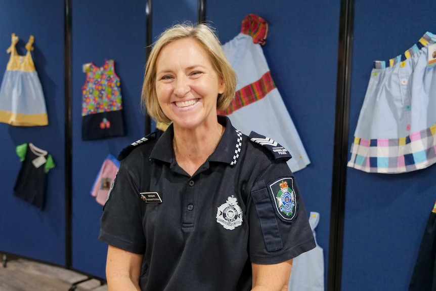 A police officer with short blonde hair smiles in front of a wall where clothing is pinned.
