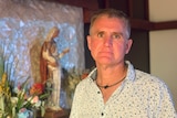 A man with grey hair stands in front of a church statue looking sad